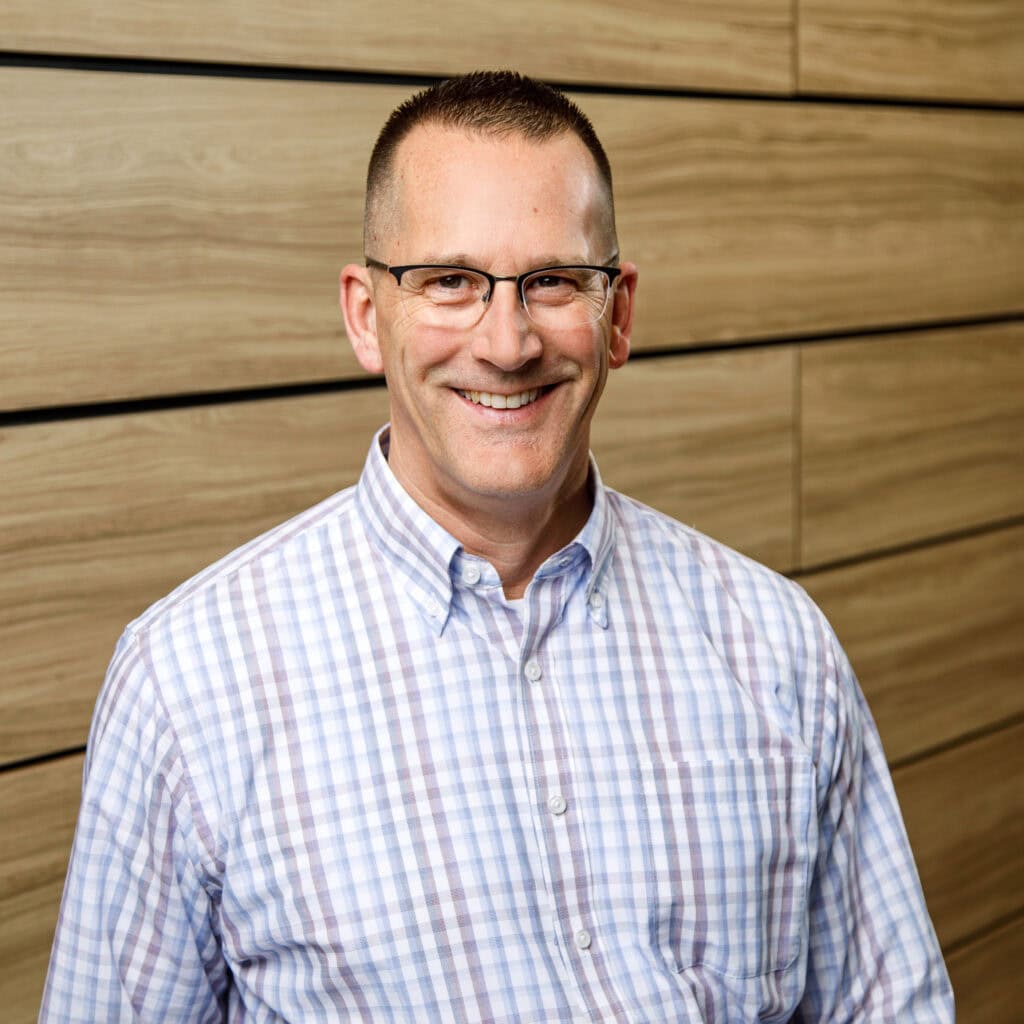A man with short hair and glasses is smiling, wearing a plaid shirt while standing in front of a wooden panel wall.
