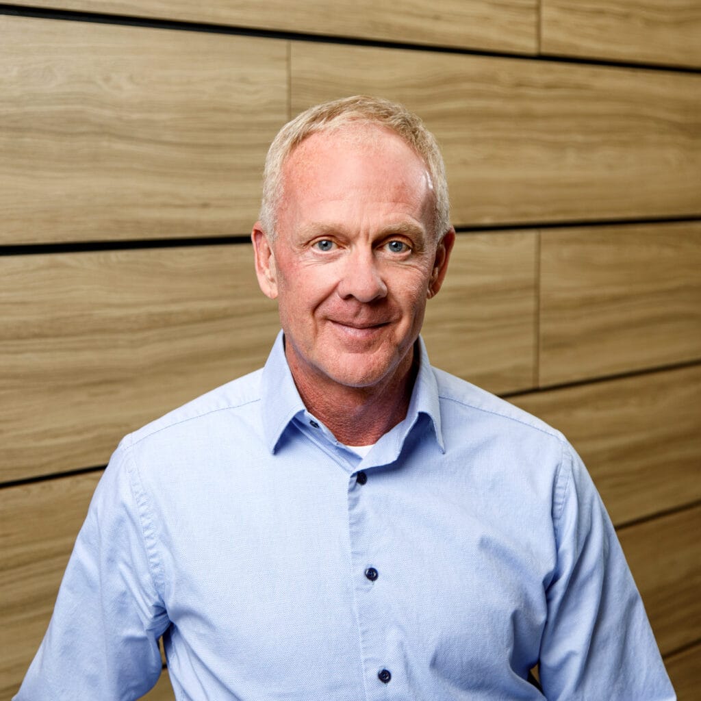 A man with short blonde hair, wearing a light blue collared shirt, standing in front of a wooden panel wall.