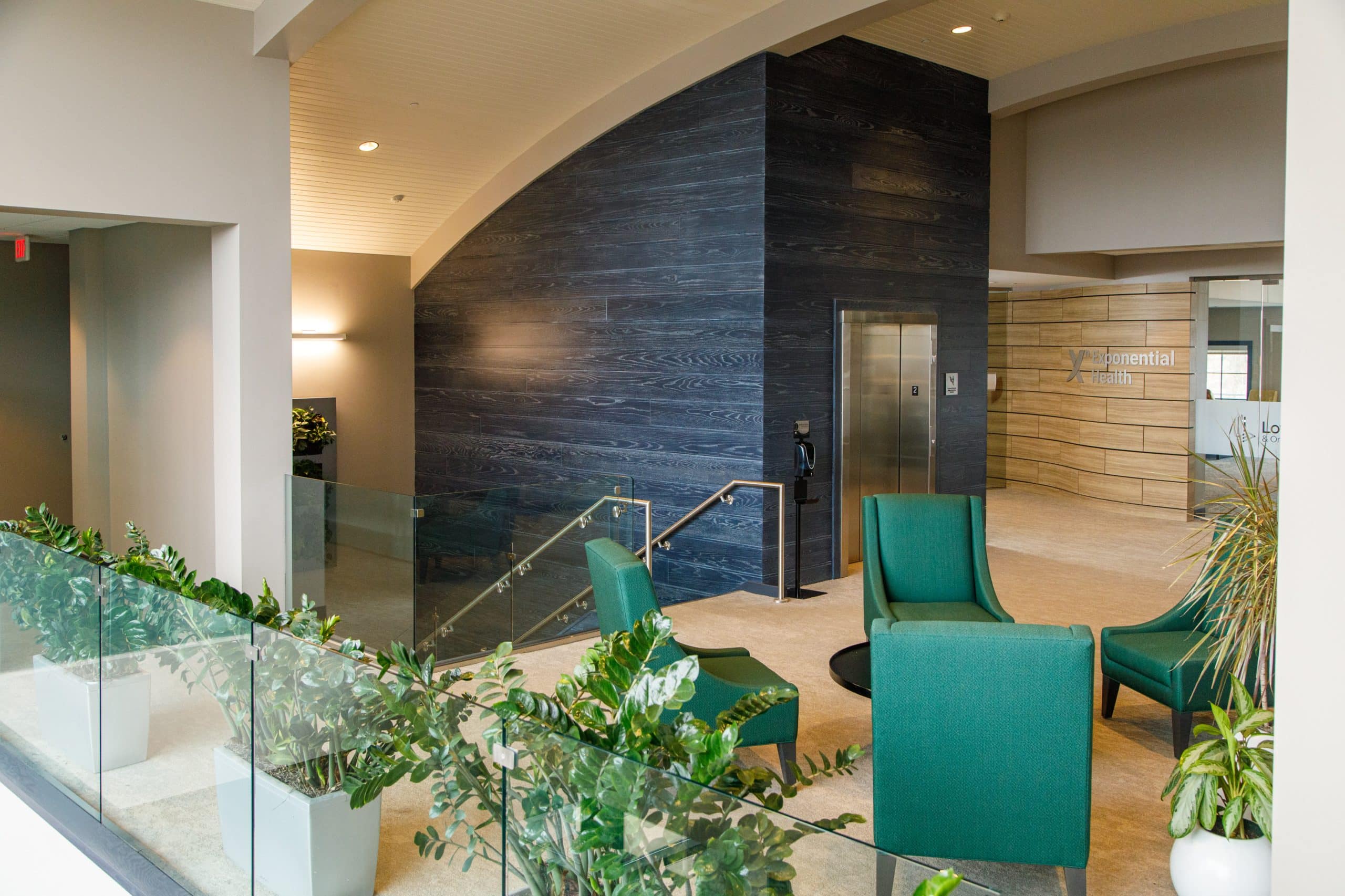 Modern office lobby with teal chairs, glass partitions, and potted plants. A wooden accent wall and an elevator are in the background.