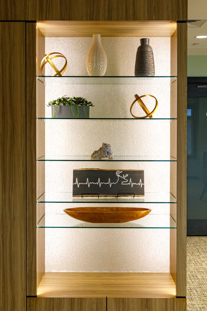 A well-lit display shelf with decorative items including abstract sculptures, vases, a small plant, a wooden bowl, and a picture of a heartbeat line with a reindeer.