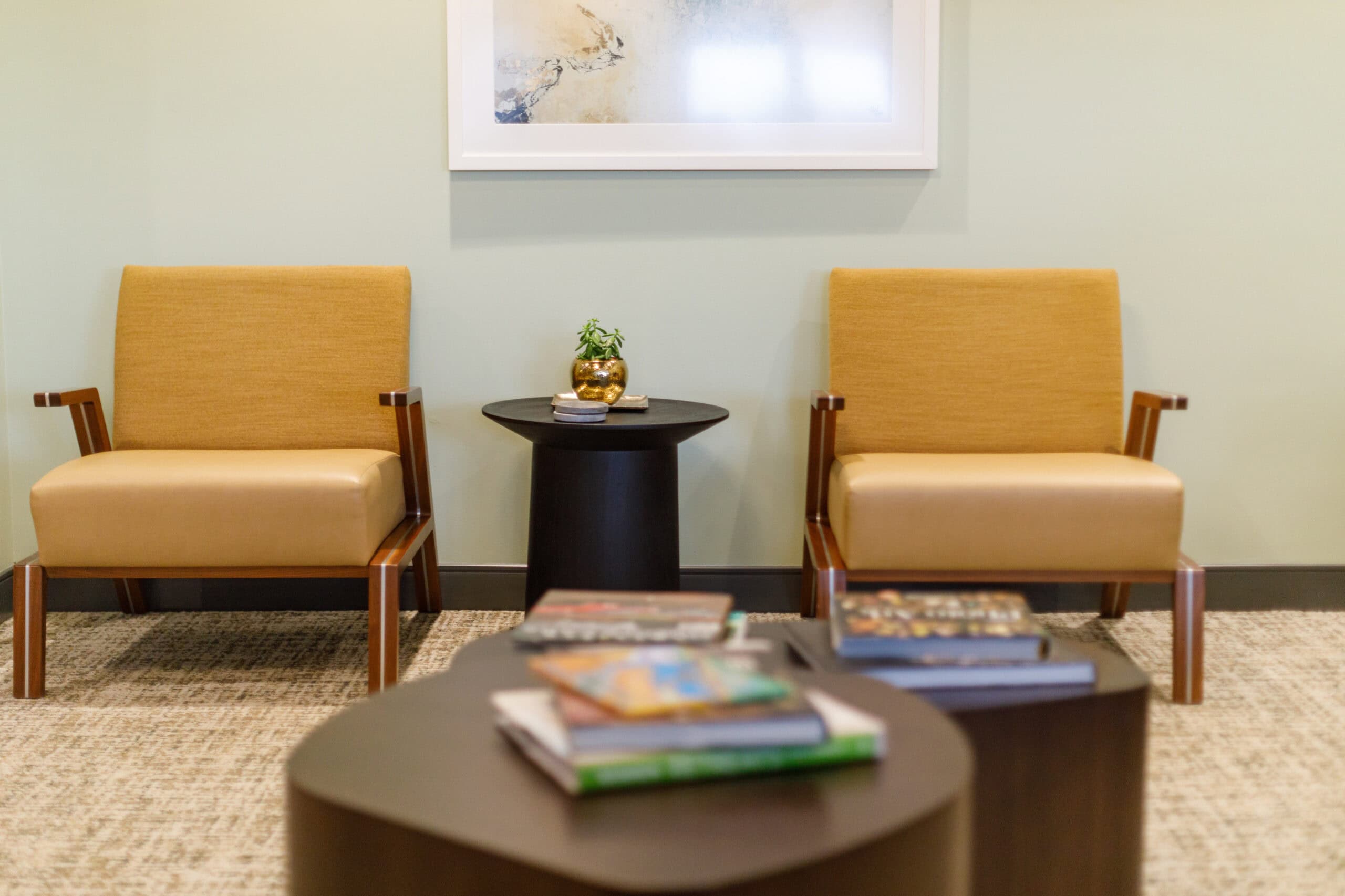 Two tan chairs with wooden arms are positioned beside a small round black table holding a small plant; books are on a larger table in the foreground.