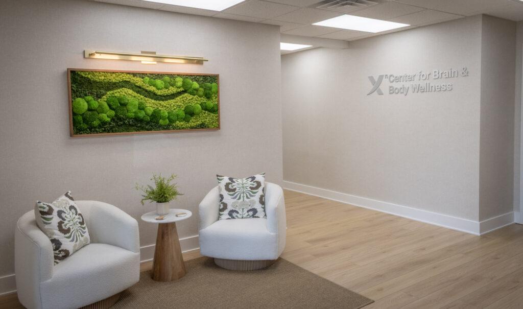 A waiting area with two white chairs, a small round table with a plant, a green moss wall art, and a sign reading "Center for Brain & Body Wellness" on a beige wall.