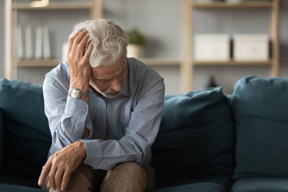 Older man sitting on a couch with his head resting on one hand, appearing thoughtful or distressed, in a living room with shelves in the background.