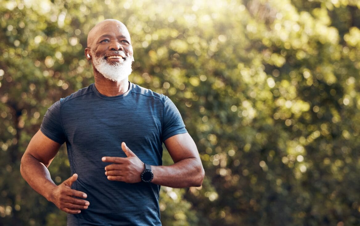 An older man with a white beard jogs outdoors, wearing a dark athletic shirt and smartwatch, with trees and sunlight in the background.