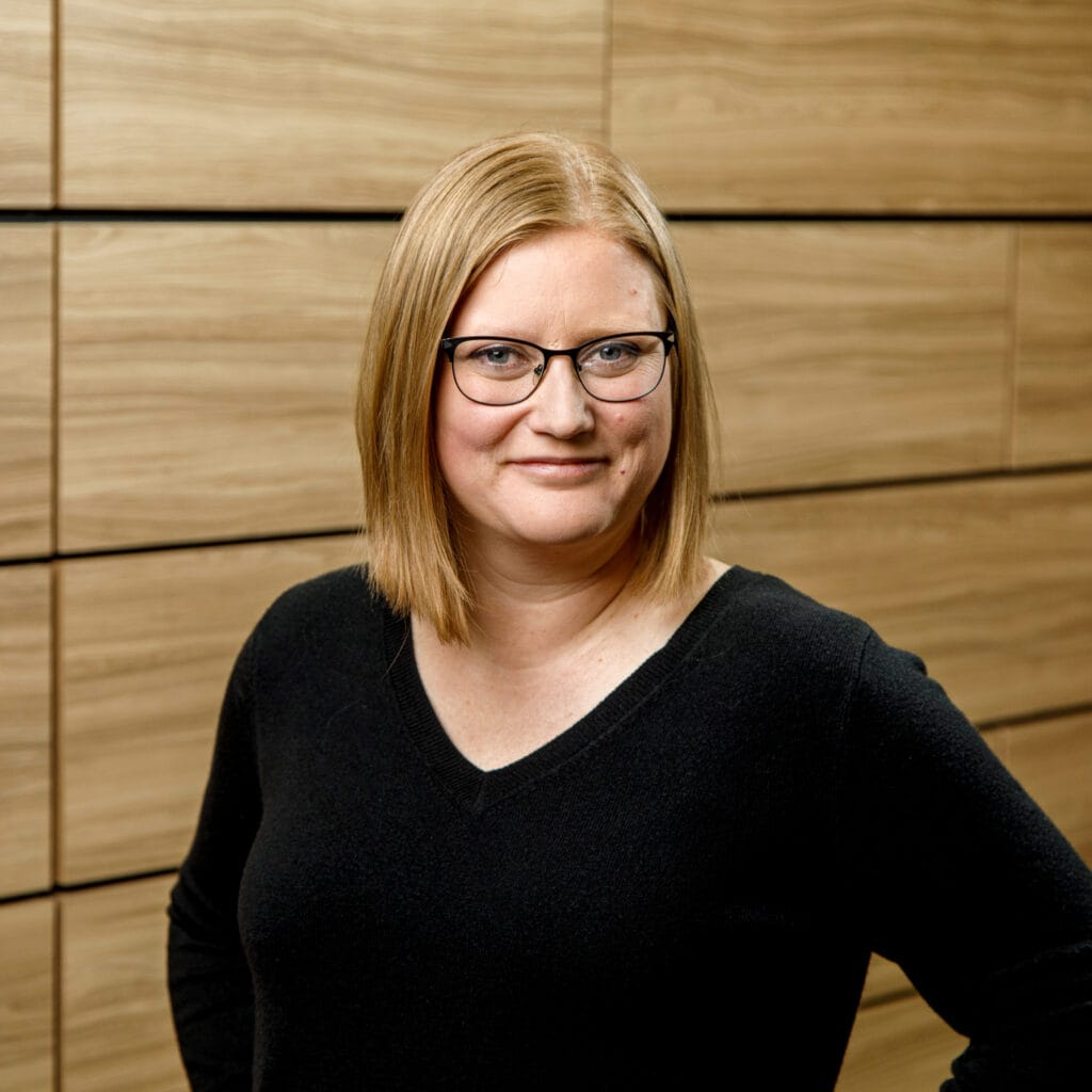 A person with glasses and straight shoulder-length hair wearing a black top stands in front of a wood-paneled wall.