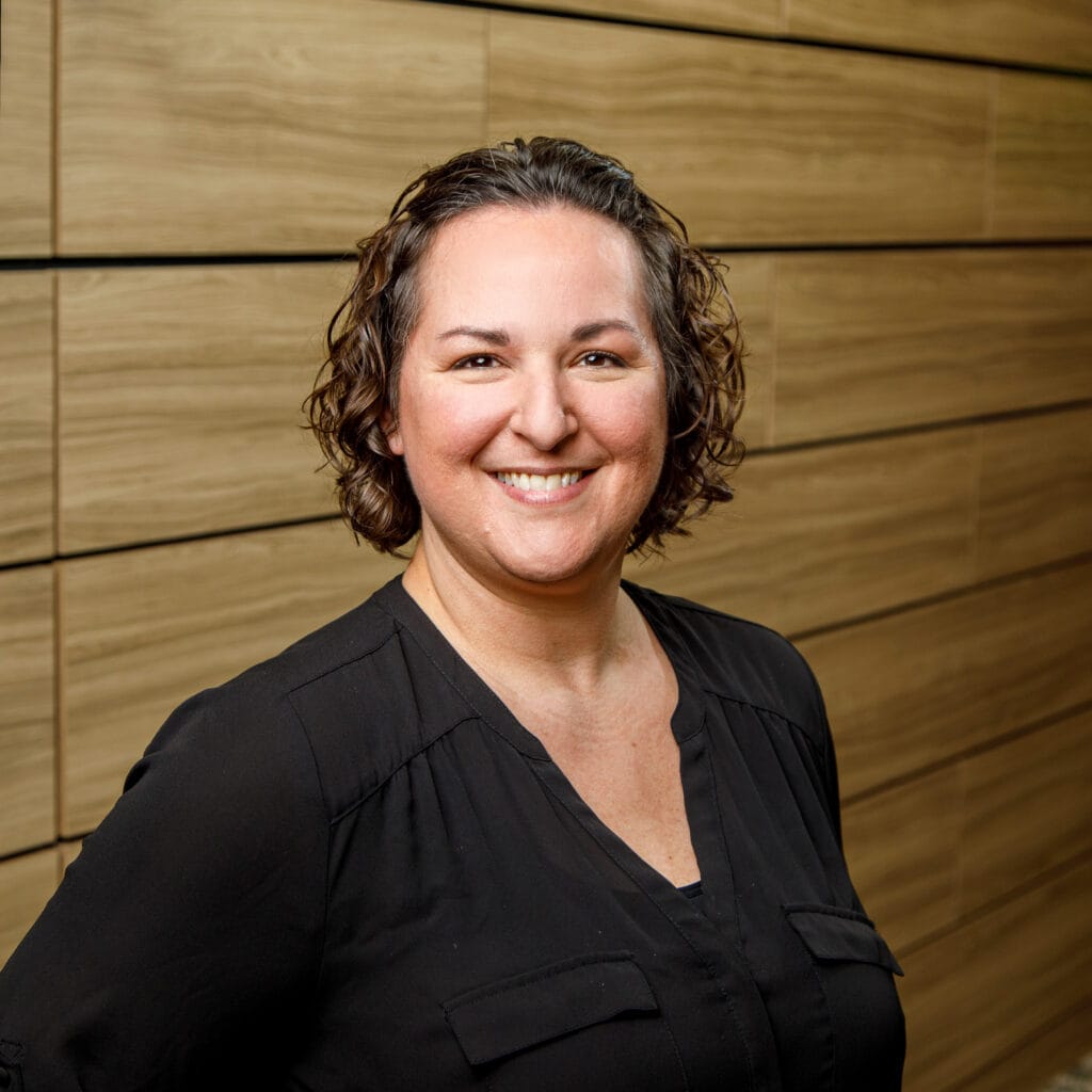 A person with short, wavy hair smiles at the camera while standing in front of a wood-paneled wall. They are wearing a black shirt.