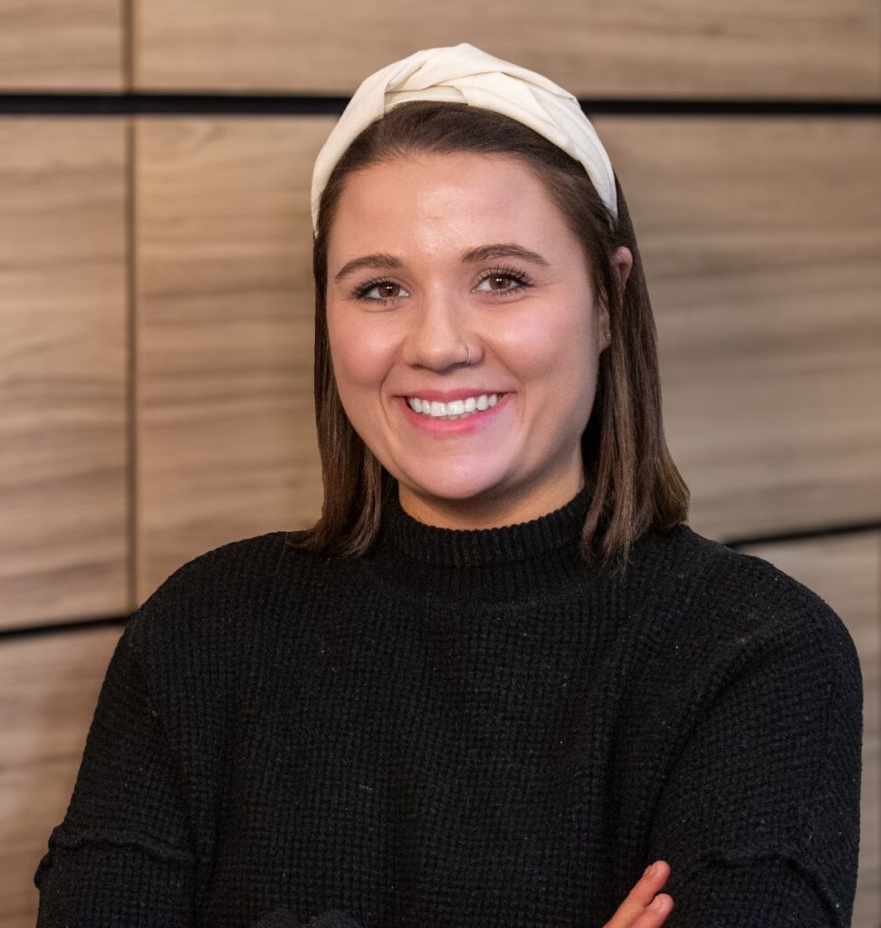 A woman with brown hair, wearing a cream headband and black sweater, smiles with arms crossed in front of a wood-paneled background.
