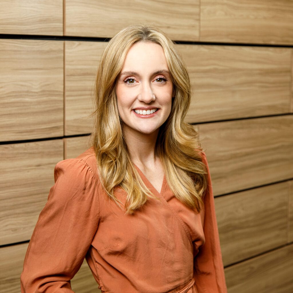 A woman with long blonde hair wearing a rust-colored blouse stands in front of a wooden panel wall, smiling at the camera.