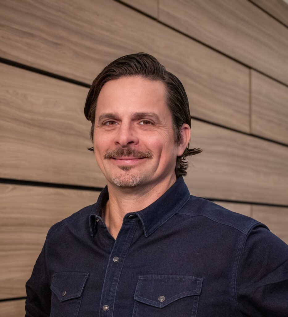 A man with dark hair and a mustache wearing a navy blue button-up shirt stands in front of a wood-paneled wall, looking at the camera and smiling slightly.