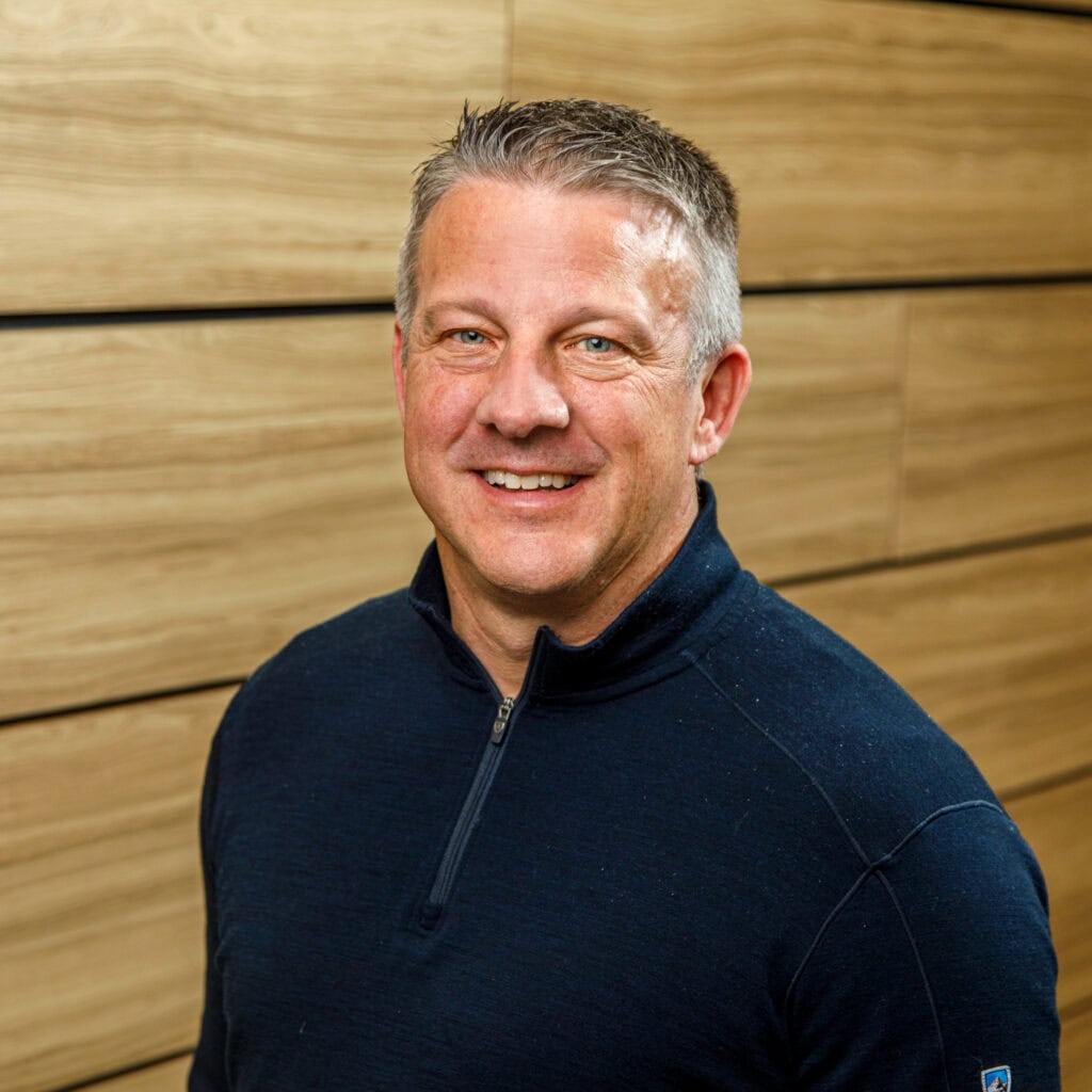 A man in a dark blue sweater stands in front of a wooden wall, smiling at the camera.