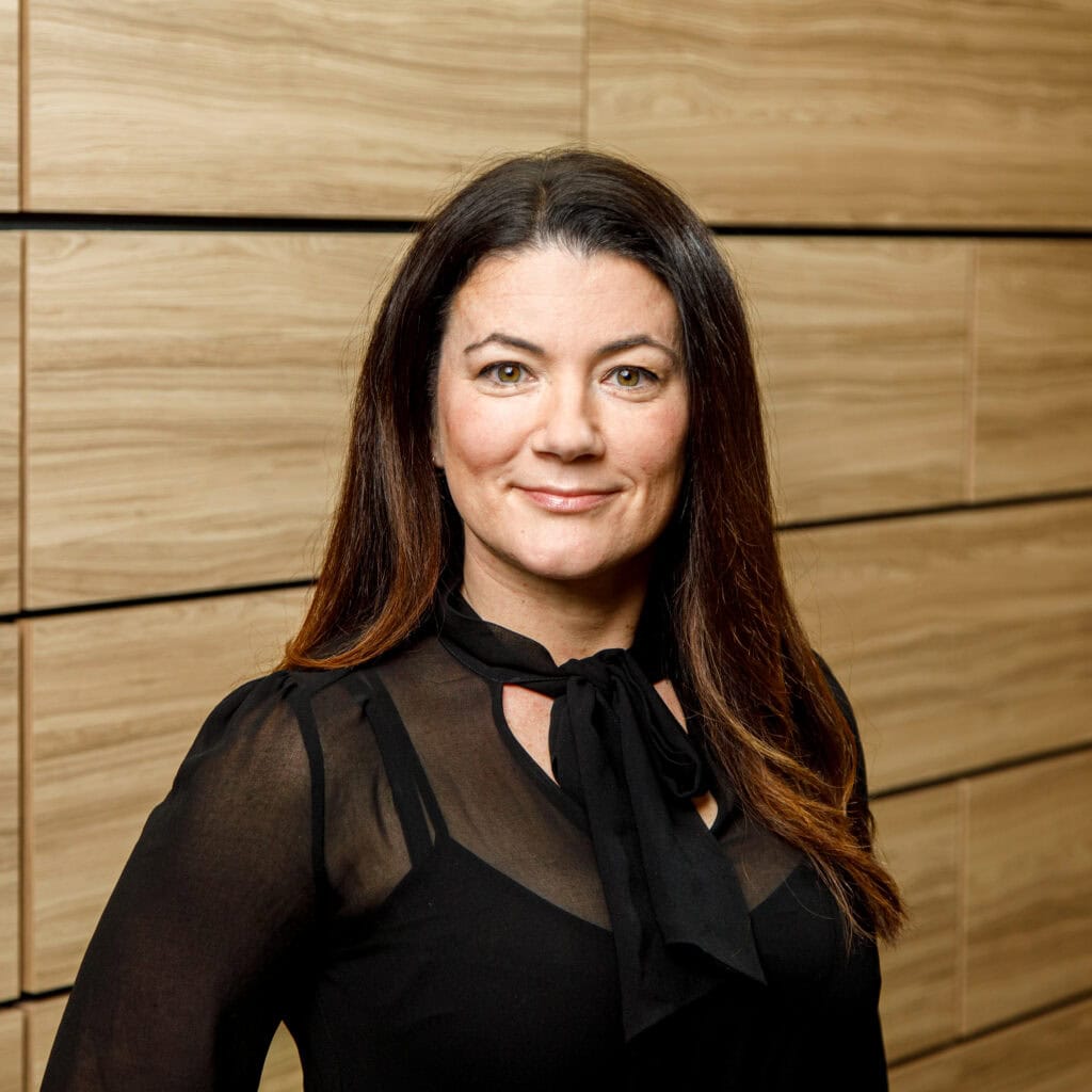 A woman with long brown hair wearing a black blouse and scarf-like neck decoration, stands in front of a wooden tiled wall.