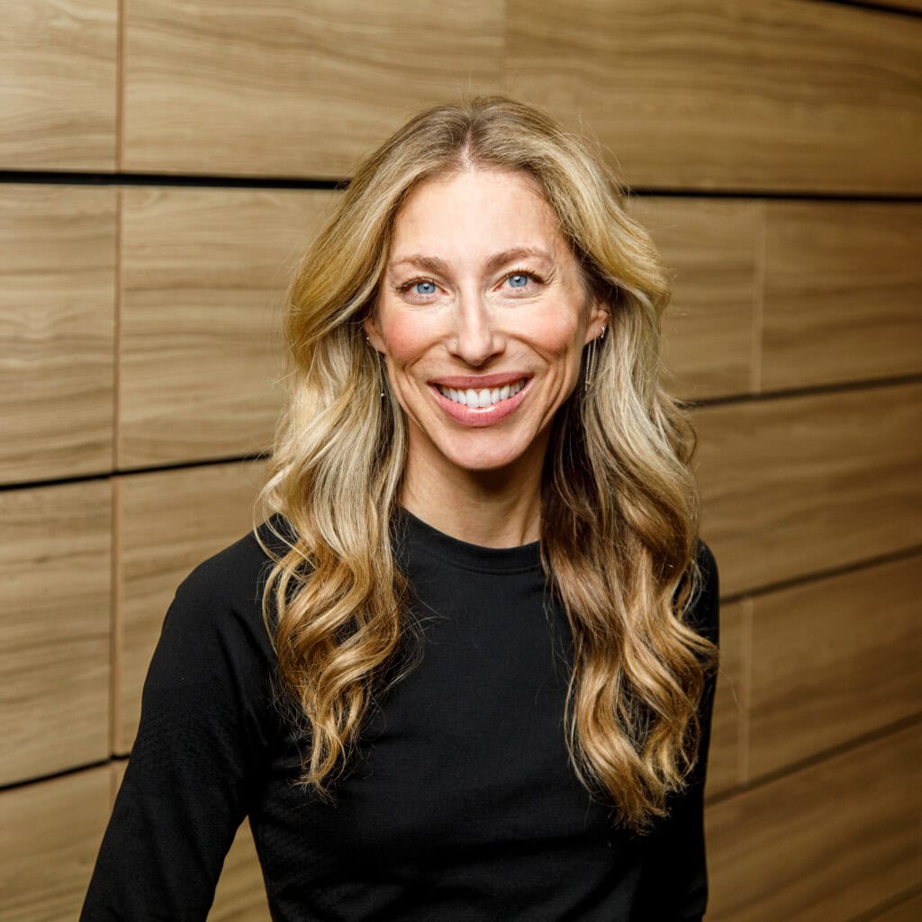 A woman with long blonde hair and a black top smiles while standing in front of a wooden wall.