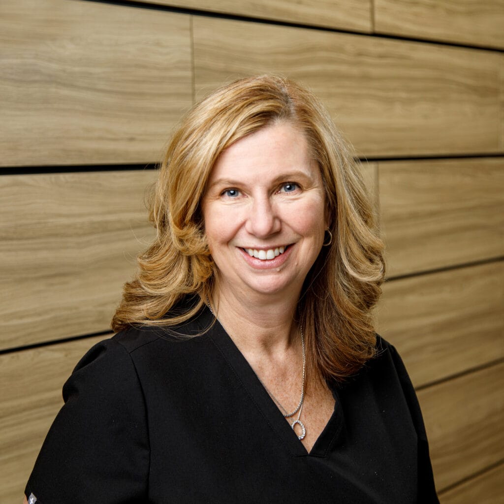A woman with shoulder-length blonde hair and wearing a black top smiles in front of a wooden wall background.