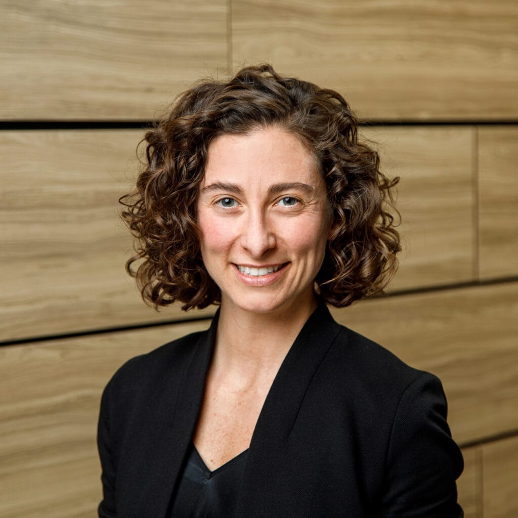 A person with curly hair, wearing a black blazer and smiling, stands in front of a wood-paneled wall.