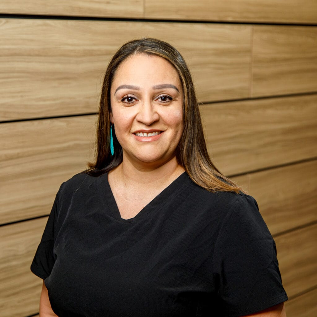 A woman with long brown hair, wearing a black top, stands in front of a wooden panel wall, smiling at the camera.