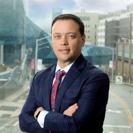 A man in a dark plaid suit and red patterned tie stands with arms crossed in front of a large window overlooking a city street.