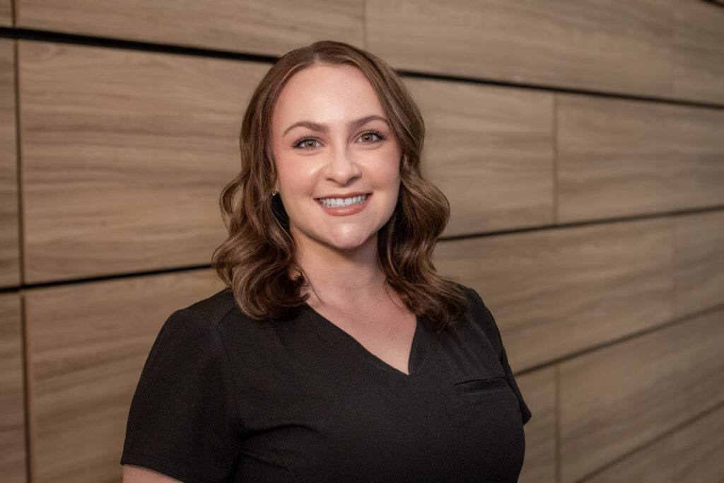 Woman with shoulder-length brown hair wearing a black top stands smiling in front of a wood-paneled wall.