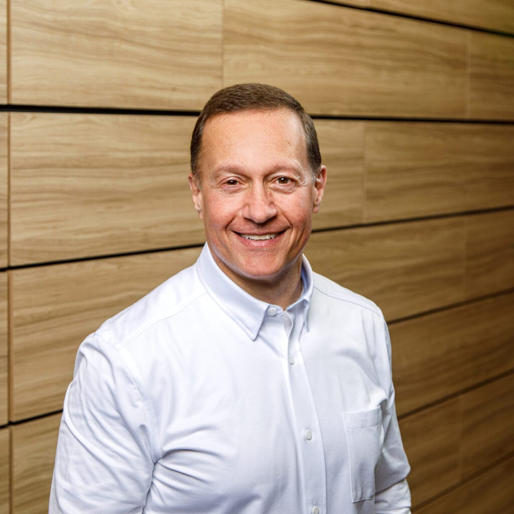 A man wearing a white button-up shirt stands smiling against a background of wooden panels.