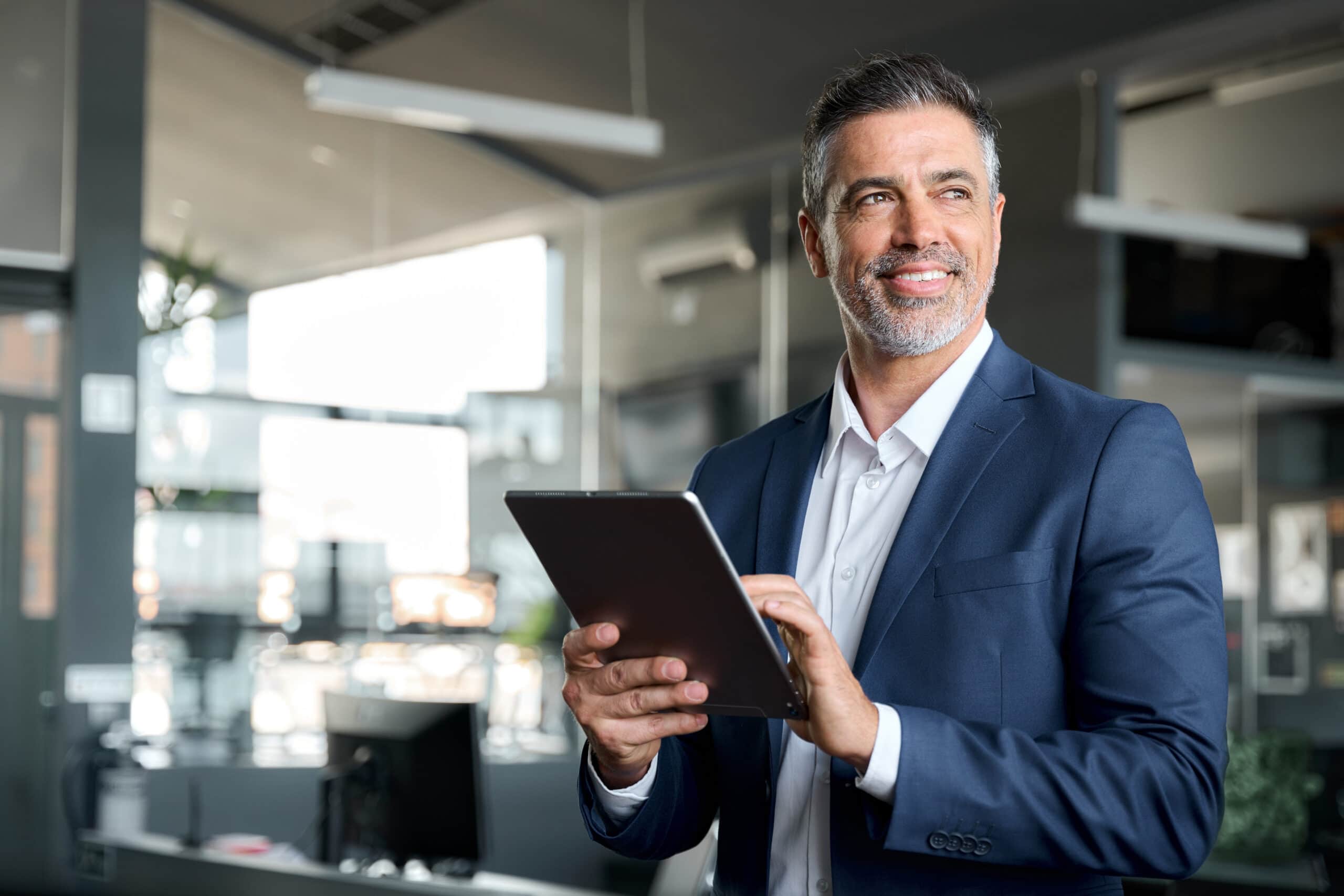 A man in a blue suit stands indoors, smiling and holding a tablet, with office furniture and large windows in the background.