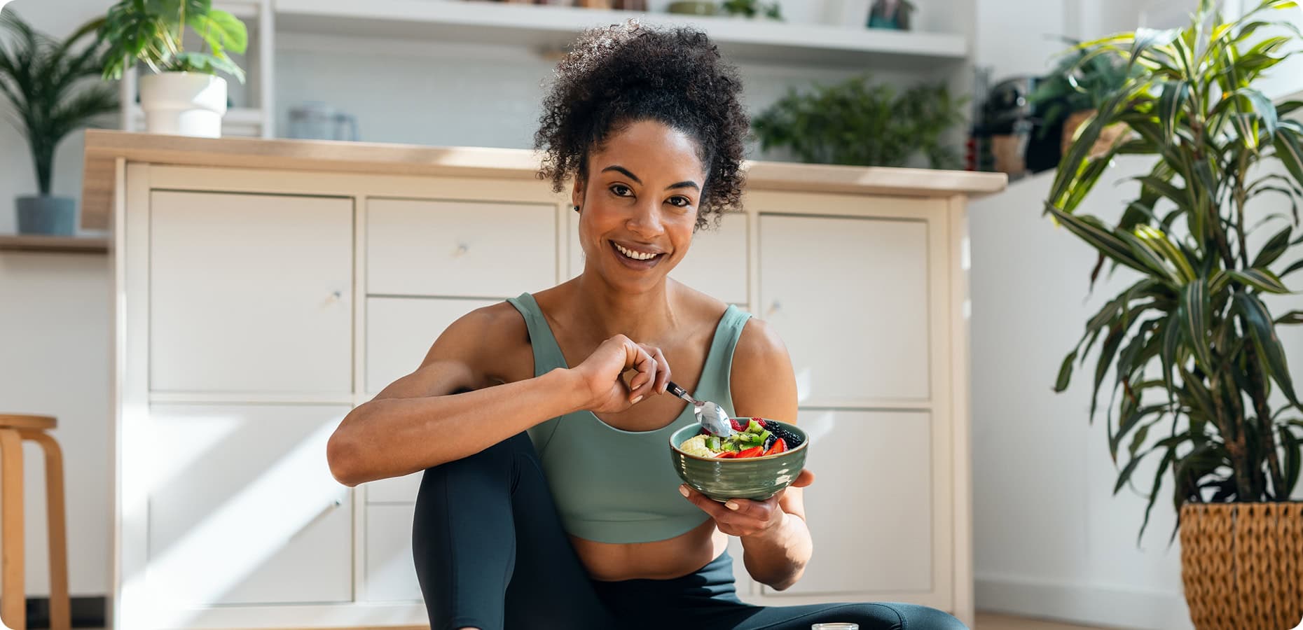 A woman in athletic wear sits on the floor in a kitchen, smiling and holding a bowl of salad with a fork.