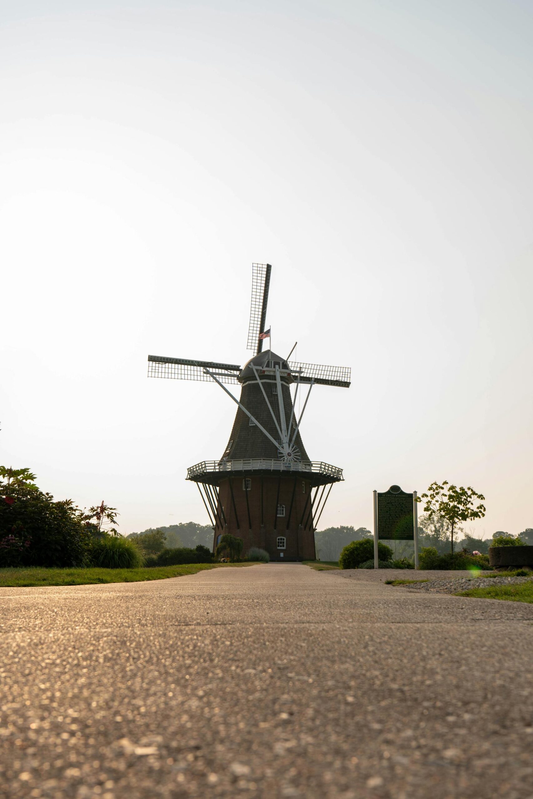 A traditional windmill stands at the end of a paved pathway, surrounded by greenery, under a clear sky.