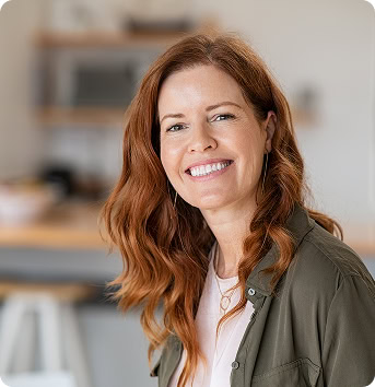 A woman with long red hair, wearing a green shirt over a light top, smiles while sitting indoors in a modern, softly focused room.