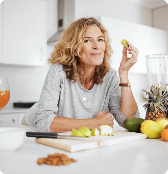 Woman standing in a kitchen, smiling while holding a piece of green apple. Chopped fruits, a knife, and a blender are on the counter.