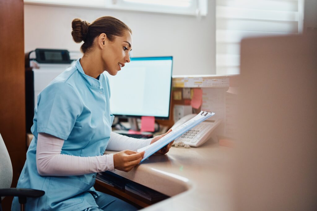 A healthcare professional in scrubs sits at a desk, reviewing documents in front of a computer and telephone in a clinical office setting.