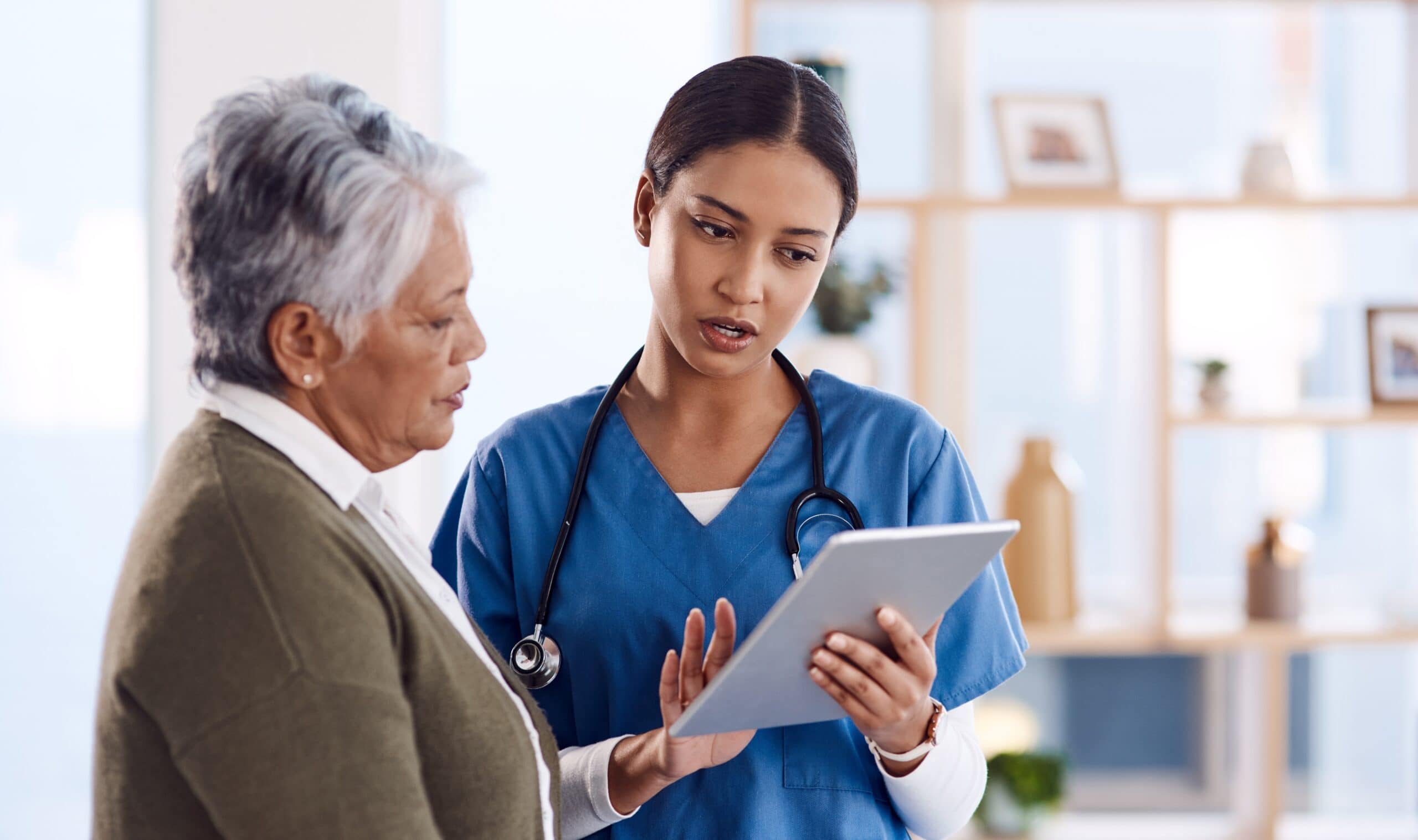 A healthcare professional in blue scrubs shows information on a tablet to an older woman in a cardigan, standing together in a well-lit room.