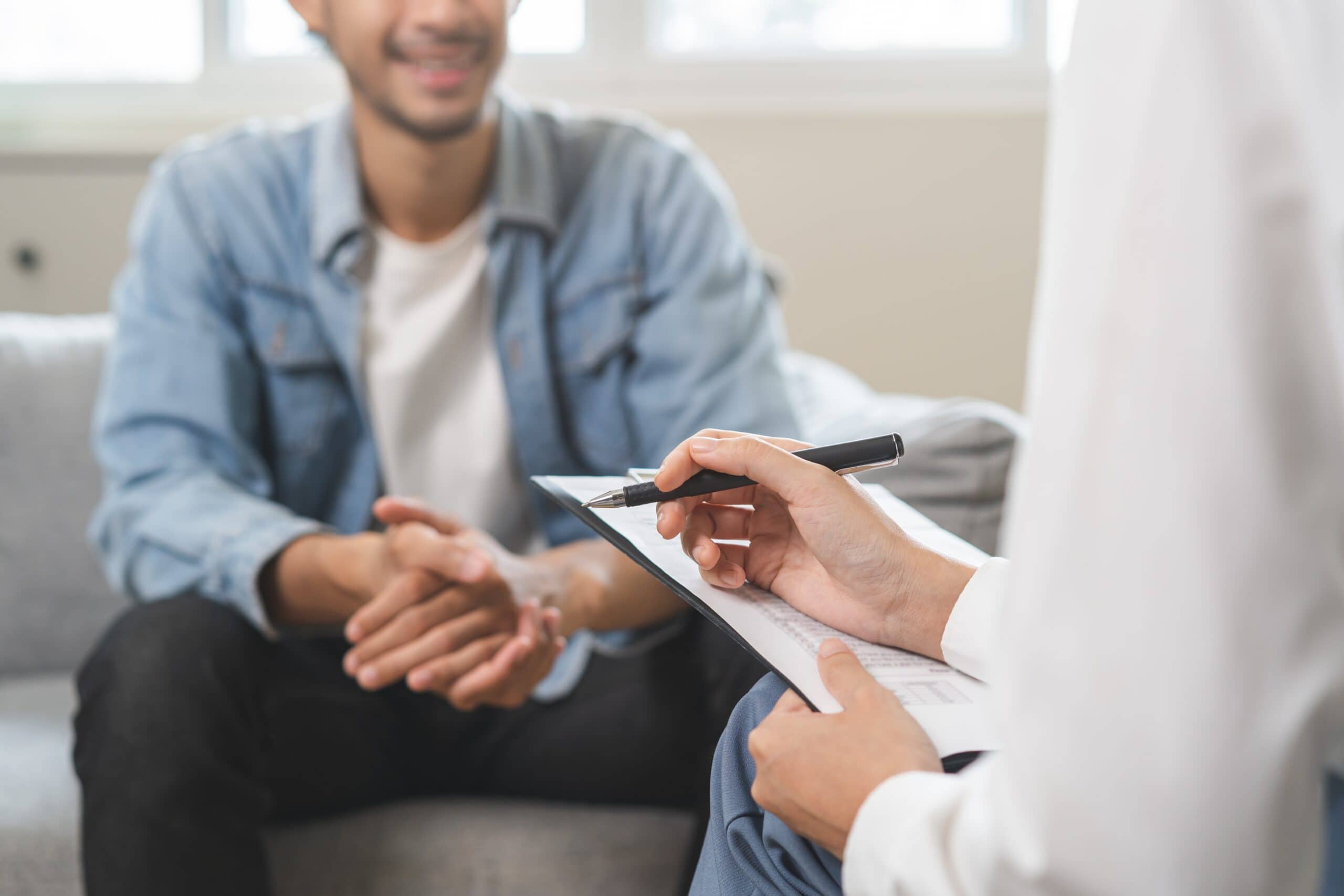 A person holding a clipboard and pen sits across from another person who is smiling and sitting on a couch, suggesting a conversation or counseling session.