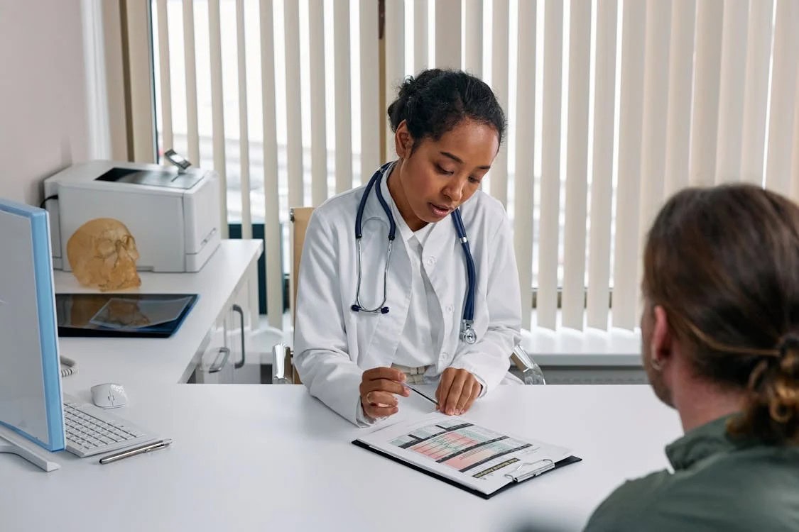 A doctor in a white coat discusses a medical chart with a patient across a desk in a well-lit office.