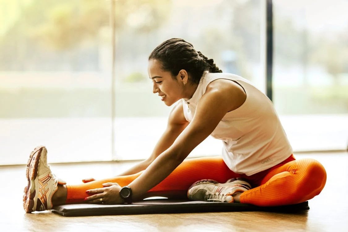A woman in athletic wear sits on a mat indoors, stretching her leg and reaching for her foot with one hand.